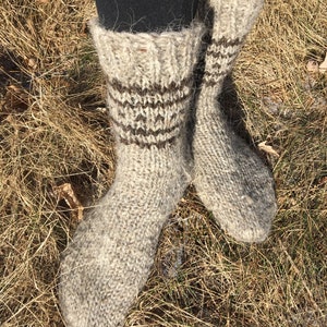 May include: A pair of thick, hand-knitted wool socks in a light brown color with brown stripes around the ankle. The socks are worn and are sitting on a bed of dry grass.