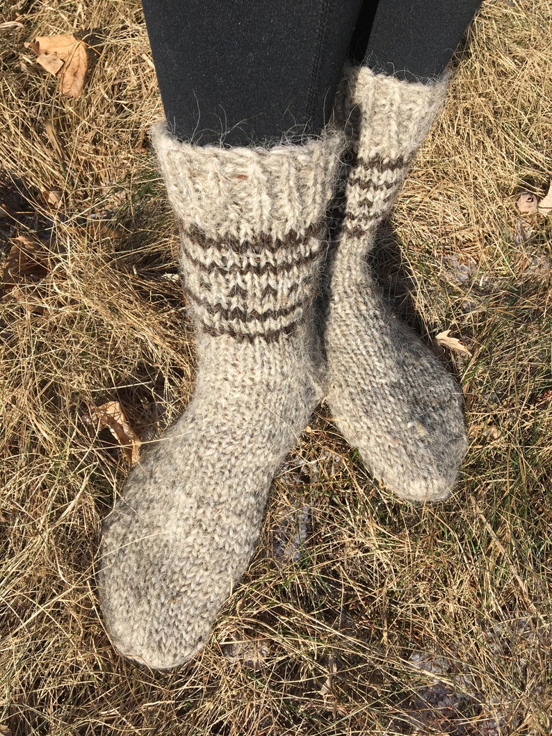 May include: A pair of thick, hand-knitted wool socks in a light brown color with brown stripes around the ankle. The socks are worn and are sitting on a bed of dry grass.