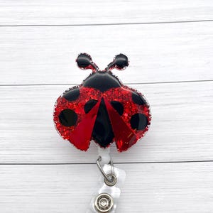 May include: Red and black glitter ladybug charm with black dots on the wings attached to a clear plastic retractable badge holder.