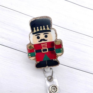 May include: A red and gold glitter nutcracker charm with a black hat and a white face. The charm is attached to a clear plastic badge holder with a silver clip.