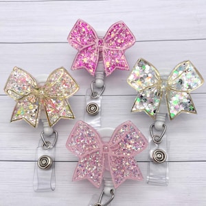 May include: Three sparkly glitter bow badge reels with clear plastic clips. The bows are pink, white and gold.