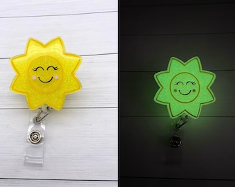 May include: A yellow glitter sun with a smiling face and closed eyes on a retractable badge reel. The sun glows green in the dark.
