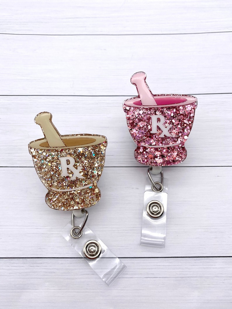 May include: Two glitter-covered mortar and pestle badge reels with a white "Rx" on the front. One is pink glitter and the other is gold glitter.