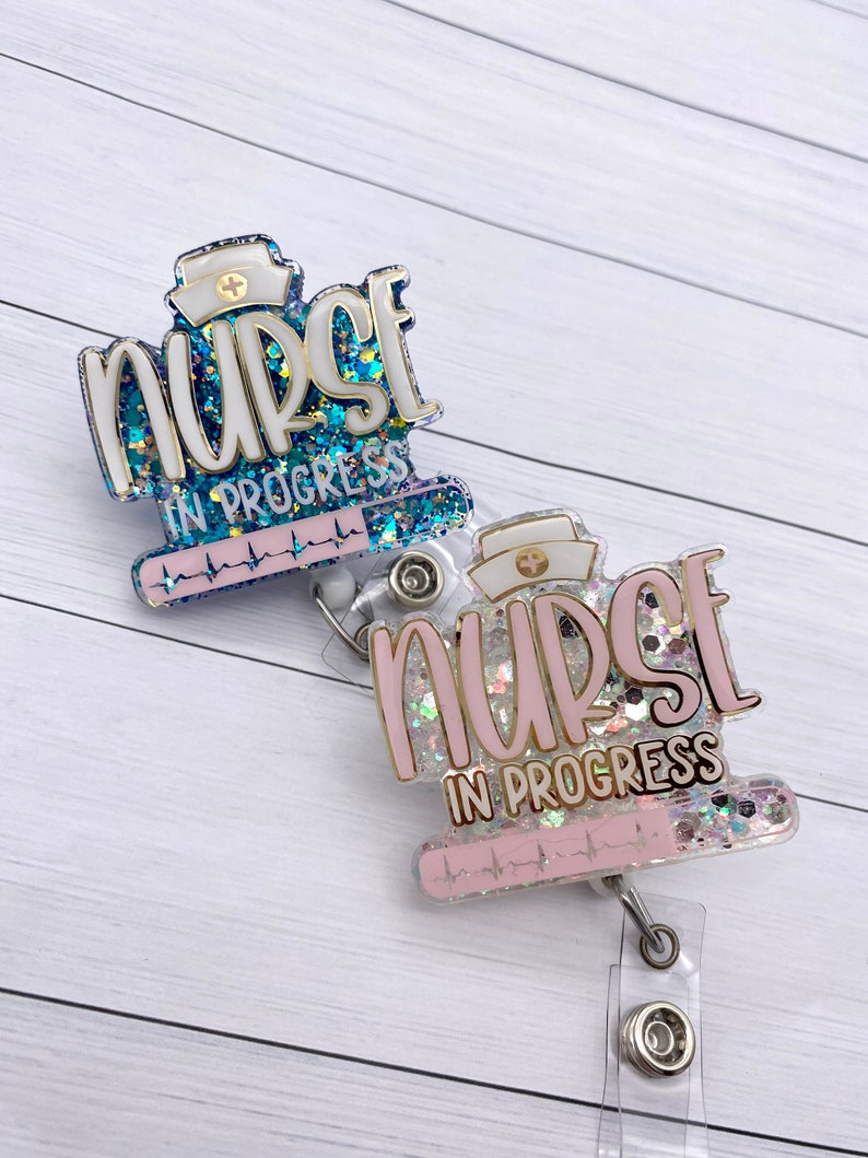 May include: Two glitter nurse badge reels with a white background. The top reel has a blue glitter background with the text "nurse in progress" and a heart rate line. The bottom reel has a pink glitter background with the text "nurse in progress" and a heart rate line.