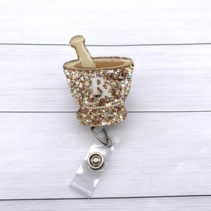 May include: Gold glitter mortar and pestle with a white "Rx" on the side, attached to a clear plastic badge holder with a silver clip.