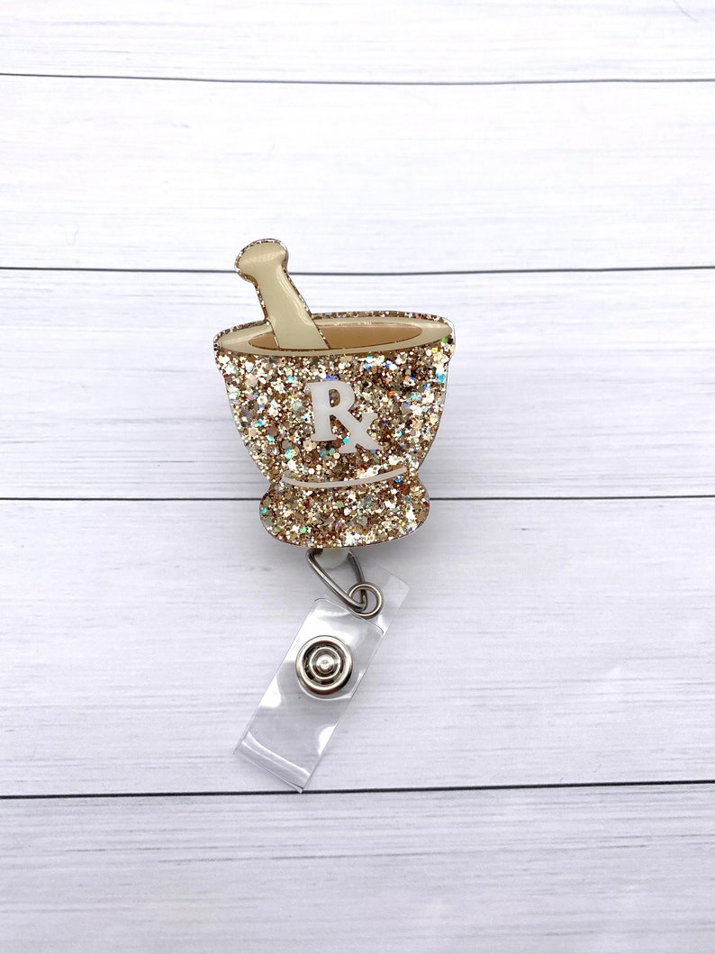 May include: Gold glitter mortar and pestle with a white "Rx" on the side, attached to a clear plastic badge holder with a silver clip.