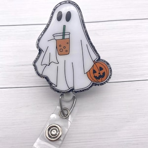 Puede incluir: Un portatarjetas con tem&aacute;tica de Halloween con un fantasma blanco sosteniendo una bebida y una calabaza. El fantasma tiene ojos negros y un contorno negro con purpurina. El portatarjetas tiene una correa de pl&aacute;stico transparente y un clip de metal.