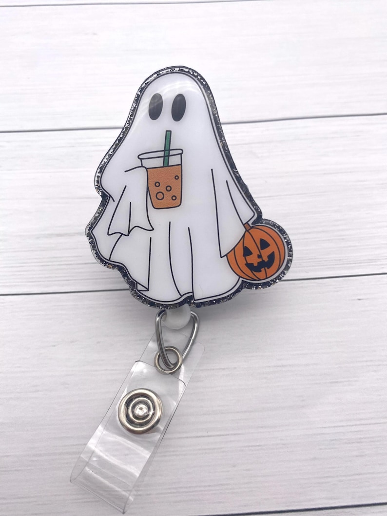 Puede incluir: Un portatarjetas con tem&aacute;tica de Halloween con un fantasma blanco sosteniendo una bebida y una calabaza. El fantasma tiene ojos negros y un contorno negro con purpurina. El portatarjetas tiene una correa de pl&aacute;stico transparente y un clip de metal.