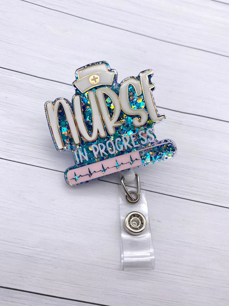 May include: A blue and white glitter badge holder with the text "Nurse in Progress" and a heart rate line. The badge holder has a clear plastic retractable clip.