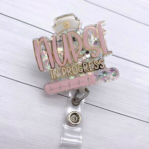May include: Pink and gold glitter badge holder with the text "NURSE IN PROGRESS" and a heart rate line. The badge holder has a clear plastic clip.
