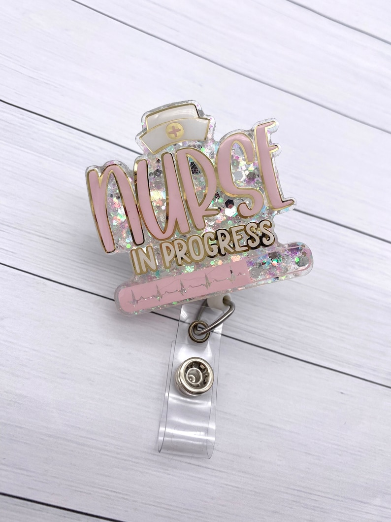May include: Pink and gold glitter badge holder with the text "NURSE IN PROGRESS" and a heart rate line. The badge holder has a clear plastic clip.