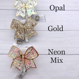 K&ouml;nnte beinhalten: Drei glitzernde Schleifen-Badge-Rollen mit durchsichtigen Plastikb&auml;ndern. Die obere Rolle ist in Opal-Glitter, die mittlere in Gold-Glitter und die untere in Neon-Mix-Glitter.