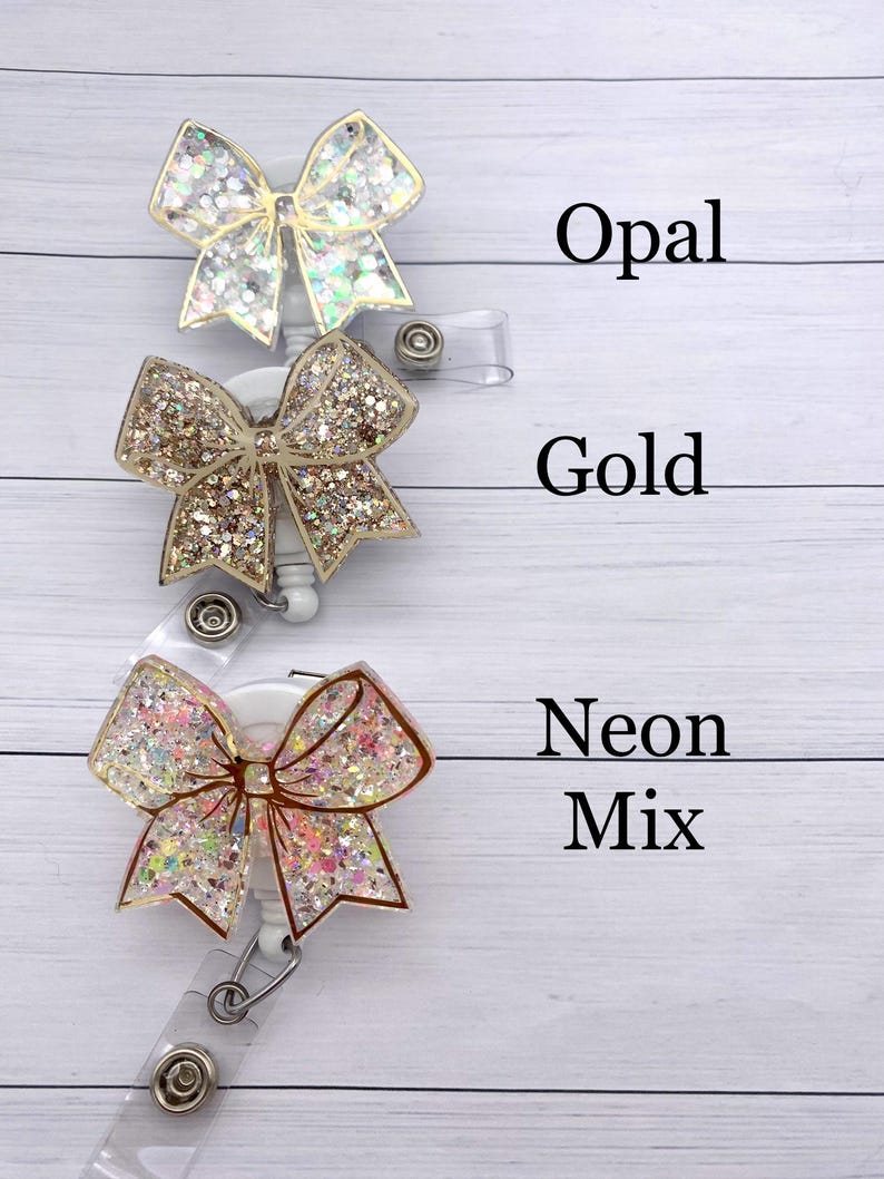 K&ouml;nnte beinhalten: Drei glitzernde Schleifen-Badge-Rollen mit durchsichtigen Plastikb&auml;ndern. Die obere Rolle ist in Opal-Glitter, die mittlere in Gold-Glitter und die untere in Neon-Mix-Glitter.