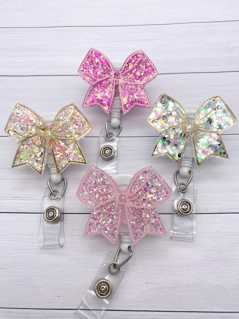 K&ouml;nnte beinhalten: Drei glitzernde Schleifen-Badge-Rollen mit durchsichtigen Plastikclips. Die Schleifen sind rosa, wei&szlig; und gold.