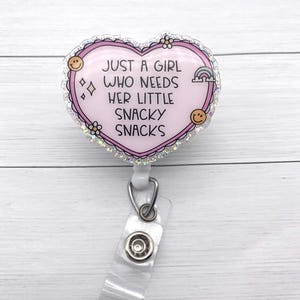 May include: A pink heart-shaped badge reel with the text "JUST A GIRL WHO NEEDS HER LITTLE SNACKY SNACKS." The badge reel has a glittery border and features small illustrations of a rainbow, smiley faces, and flowers.
