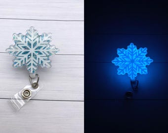 Può includere: Un portabadge a forma di fiocco di neve bianco con dettagli di glitter blu. Il portabadge brilla di blu al buio.