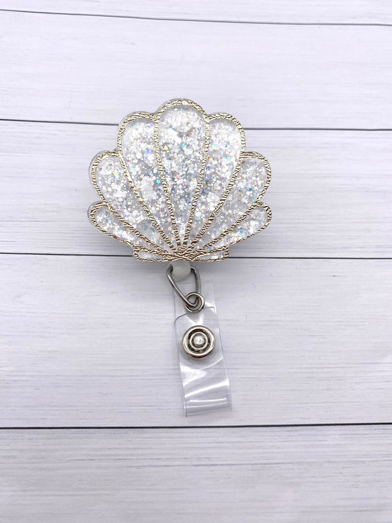 May include: A white glitter seashell with gold accents attached to a clear plastic retractable badge holder.