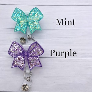 K&ouml;nnte beinhalten: Zwei glitzernde Schleifen-Ausweishalter mit Ausziehm&ouml;glichkeit. Der obere ist mintgr&uuml;n und der untere ist lila. Der Text "Mint" und "Purple" steht unter jeder Schleife.