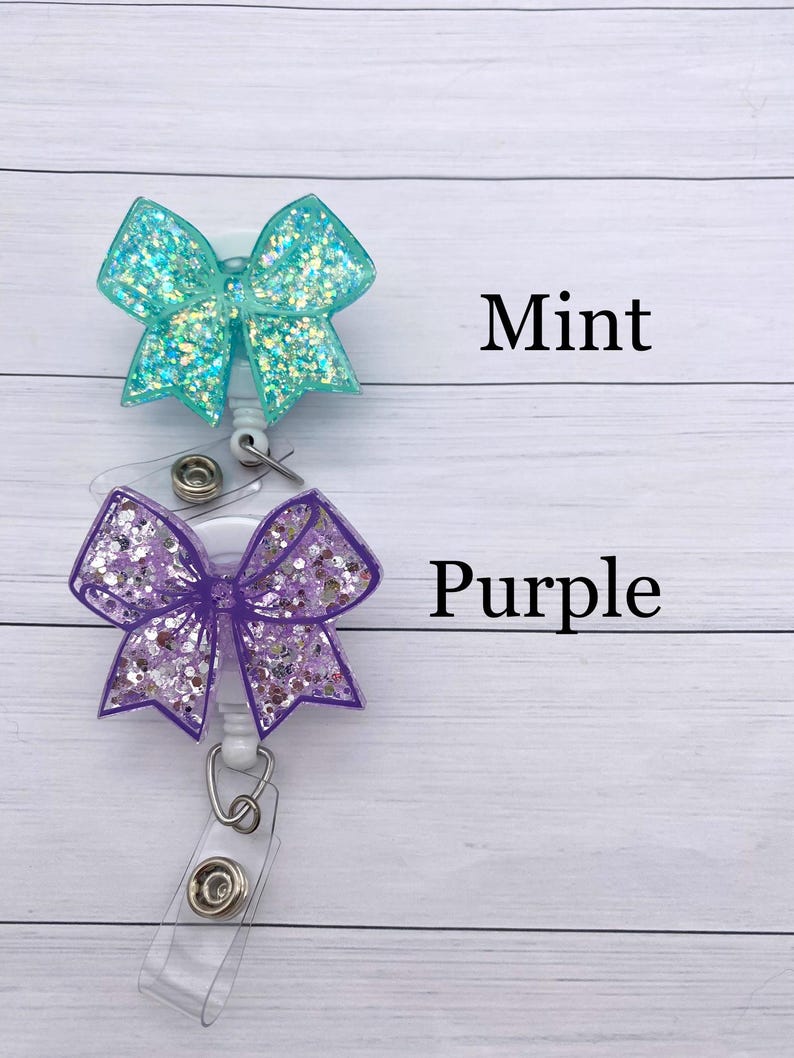 K&ouml;nnte beinhalten: Zwei glitzernde Schleifen-Ausweishalter mit Ausziehm&ouml;glichkeit. Der obere ist mintgr&uuml;n und der untere ist lila. Der Text "Mint" und "Purple" steht unter jeder Schleife.