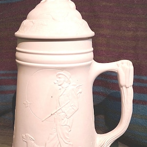 May include: White ceramic beer stein with a dog-shaped lid. The stein features a handle and a detailed relief of a person with a dog, and a hunter with a pipe. The stein is approximately 10 inches tall.