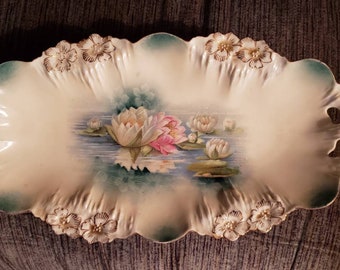 May include: An oval, off-white ceramic serving platter with a scalloped edge and floral accents. The center features a painted scene of water lilies in shades of pink, white, and green. The handles have cut-out details.