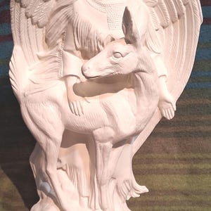 May include: A white ceramic sculpture of a woman with large feathered wings, seated on a deer. The woman wears a headpiece and fringed clothing. The sculpture is on a round base.