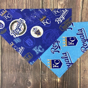 May include: Two blue and light blue dog bandanas with a Kansas City Royals theme. The bandanas feature the team's logo, the words "Royals" and "KC", and a baseball.