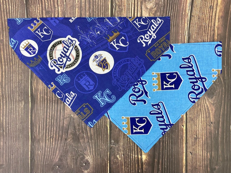 May include: Two blue and light blue dog bandanas with a Kansas City Royals theme. The bandanas feature the team's logo, the words "Royals" and "KC", and a baseball.