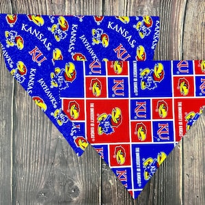May include: Two triangular pet bandanas featuring University of Kansas branding. One bandana has a blue background with repeating Jayhawk logos and KU text. The other features a red and blue grid pattern with Jayhawk logos and text.