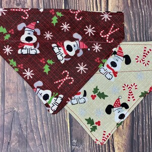 May include: Two triangular pet bandanas featuring a Christmas-themed pattern. One bandana is dark red with a plaid texture, while the other is light beige. Both display repeating illustrations of white dogs wearing red Santa hats, green holly, and candy canes.