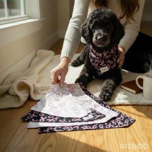 A person is holding a black dog wearing a pink ribbon bandana, with several pink ribbon-patterned dog towels or blankets on the floor in front of them.