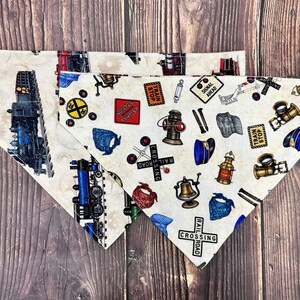 May include: Two triangular pet bandanas featuring a cream-colored background with a repeating pattern of vintage trains, railroad crossing signs, lanterns, whistles, and conductor hats. The prints include text such as Train Stop and Railroad Crossing.