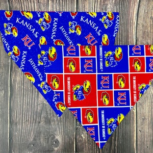 May include: Two triangular dog bandanas with a Kansas Jayhawks theme. One bandana is blue with a white KU logo and the words "Kansas Jayhawks" repeated. The other bandana is red with a white KU logo and the words "The University of Kansas" repeated.