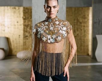 May include: A high-neck capelet featuring intricate gold beadwork and white floral appliqués, finished with long, cascading gold fringe that extends over a sheer bodice. The garment is paired with solid black trousers, creating a structured silhouette.