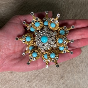 May include: A gold-tone brooch featuring a central turquoise cabochon surrounded by clear rhinestones. The outer edge consists of radiating gold-tone petals, each tipped with a clear rhinestone and accented with a round turquoise cabochon stone.