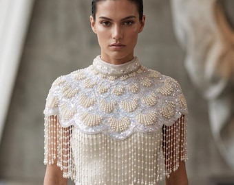 May include: A white high-neck capelet featuring intricate scalloped embroidery, embellished with rows of cream-colored pearls and shimmering sequins. The garment is finished with a long, delicate fringe of hanging pearl strands along the hem.