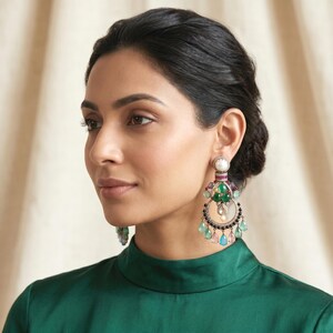 May include: A pair of ornate chandelier earrings featuring a central white pearl, a horizontal band of red stones, and a cluster of green cabochons. The circular frame is accented with small clear stones and dangling teardrop-shaped gems in green, pink, and blue.
