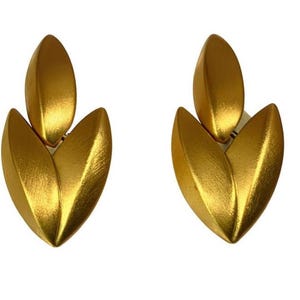 May include: A pair of gold-colored earrings. Each earring features a design of three leaf-shaped elements, with a smaller leaf above two larger ones. The earrings have a textured surface and a clip-on style.