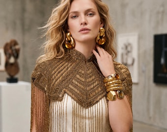 May include: A gold beaded capelet with a high collar and long fringe, worn over a white garment. The ensemble includes large gold link earrings and a stack of chunky gold bracelets featuring smooth and faceted amber-toned stones.