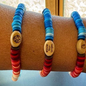 May include: Three bracelets with wooden beads and charms. The bracelets are layered on a person's arm. The charms have the text "#Show Your Stripes" and a globe with hands around it.