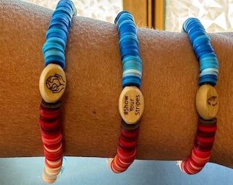 Puede incluir: Tres pulseras con cuentas y dijes de madera. Las pulseras están superpuestas en el brazo de una persona. Los dijes tienen el texto "#Show Your Stripes" y un globo terráqueo con manos alrededor.