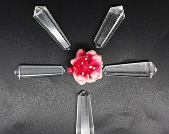 Könnte beinhalten: Fünf klare Quarzstabkristalle, angeordnet um eine rosa-rote Blume auf einer dunkelgrauen Oberfläche. Die Stäbe sind facettiert und an einem Ende spitz, mit rechteckiger Form. Die Blume hat mehrere Blütenblätter.