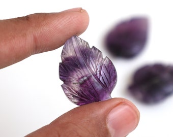 Puede incluir: Una mano sosteniendo una hoja de fluorita púrpura tallada con vetas detalladas. Dos tallas de hojas adicionales están ligeramente desenfocadas en el fondo. Las hojas son una mezcla de púrpura y blanco, sobre un fondo blanco.