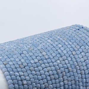 May include: Close-up of a strand of light blue gemstone beads. The beads are small, square-shaped, and faceted, creating a textured surface. The color is a soft, muted blue, and the beads are strung together, forming a continuous line.