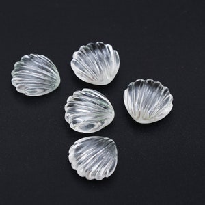 Pode incluir: Cinco contas transparentes em forma de concha com uma textura estriada dispostas sobre uma superfície preta. As contas são translúcidas, permitindo a passagem da luz, e provavelmente são feitas de vidro ou material semelhante. As conchas têm cerca de 2,5 cm de diâmetro.