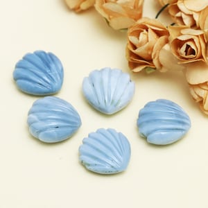 Op de afbeelding: Vijf lichtblauwe schelpvormige kralen met gedetailleerde geribbelde oppervlakken. De kralen zijn gerangschikt op een lichte achtergrond, met gedroogde bloemen in de rechterbovenhoek. Ideaal voor het maken van sieraden en knutselprojecten.