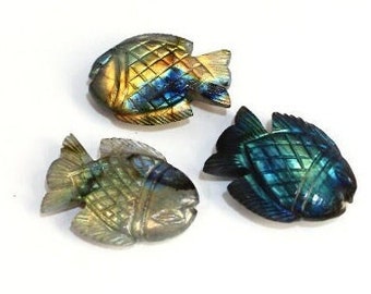 Puede incluir: Tres figuras de peces de piedra tallada en tonos dorados, azules y verdes. Cada pez tiene escamas y aletas detalladas. Las figuras están dispuestas sobre una superficie blanca.