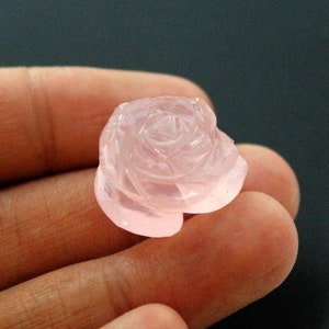 Puede incluir: Una gema tallada en forma de rosa, de un tono rosa pálido. La gema se sostiene en una mano, mostrando los intrincados detalles de los pétalos. La piedra translúcida captura la luz, resaltando la delicada talla.