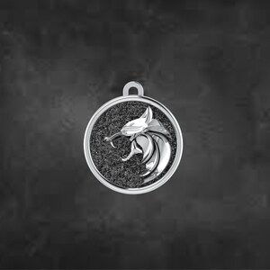 May include: A silver pendant featuring a lion's head in profile, with an open mouth, set against a textured, dark grey background within a circular frame. The pendant has a loop for a chain.