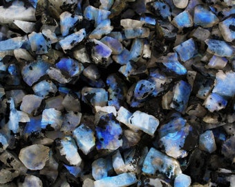 May include: A collection of raw, uncut moonstone crystals. The stones display a mix of colors, including white, blue, and black. The crystals have a rough, natural texture and are irregularly shaped.